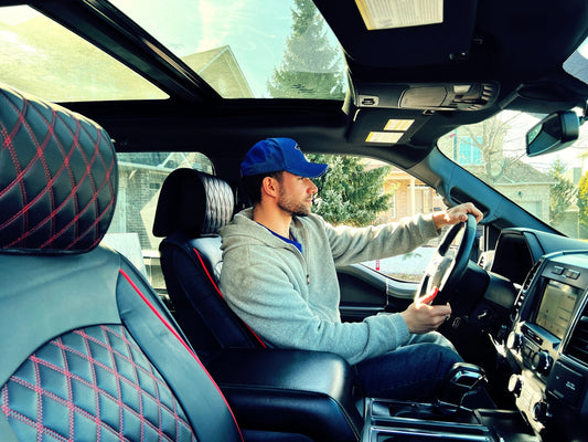 Man wearing blue cap and gray hoodie driving truck with black leather seats and sunroof on sunny day